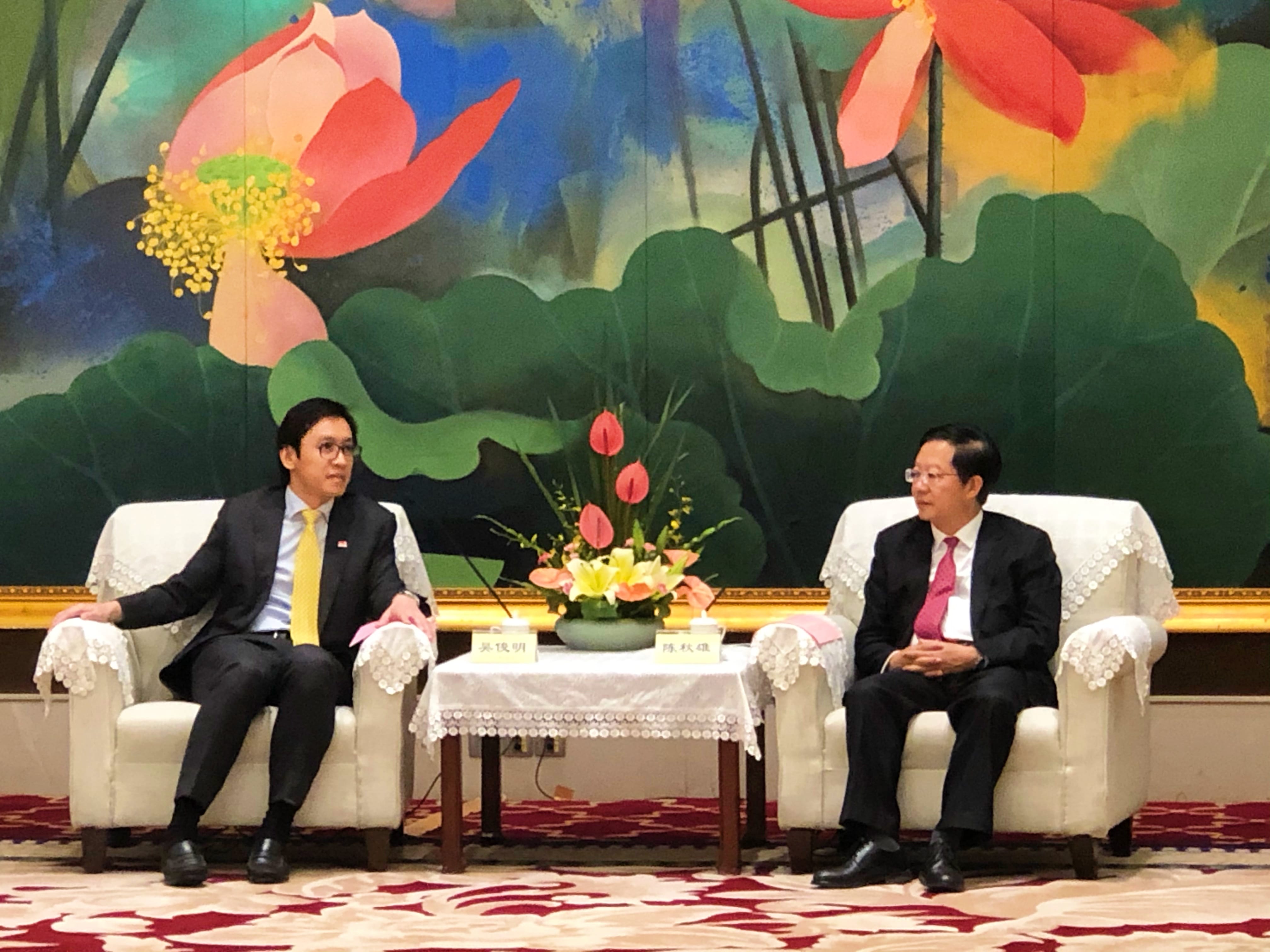 Two men in suits sit in chairs, a floral arrangement on a lace-covered table between them, lotus painting behind.